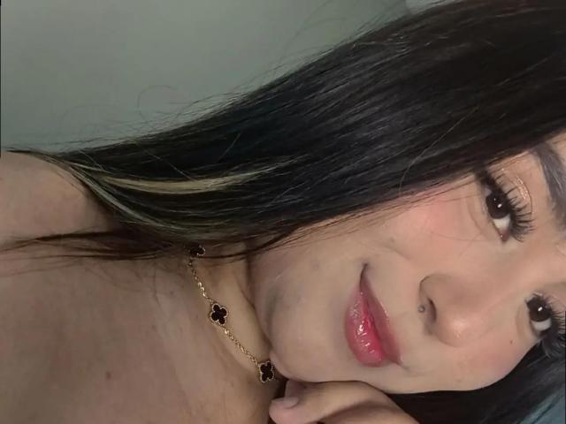 SalomettX has long black hair with blonde highlights, brown eyes, and a small nose ring. She's smiling with glossy pink lips, wearing a black flower necklace.
