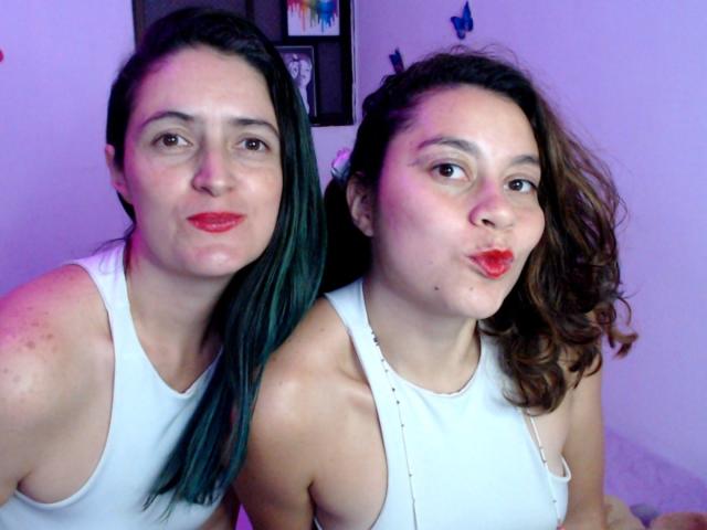 LunaVenus with green-tinted black hair and LunaVenus with brown wavy hair, both pouting red lips, wearing white tank tops, standing close, arms around each other's shoulders.