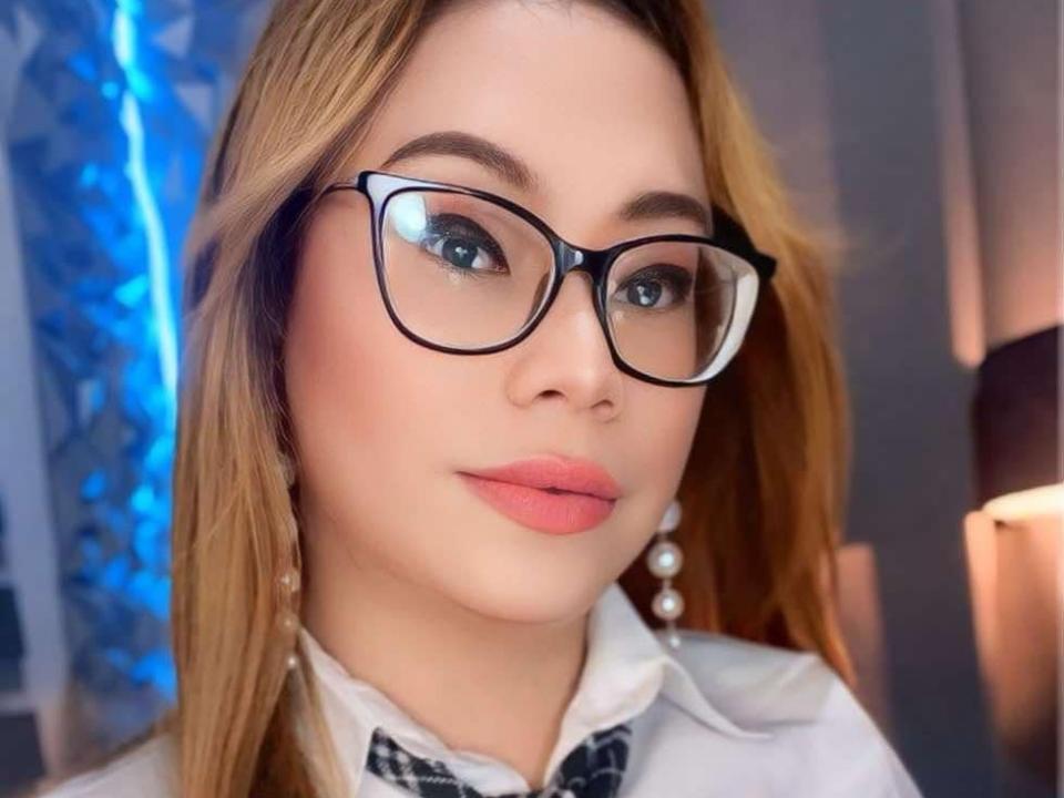 QueenStarlet, com óculos, batom rosa e brincos de pérolas, tem cabelo castanho claro e olhos azuis. Ela está vestindo uma camisa branca com um padrão xadrez preto..