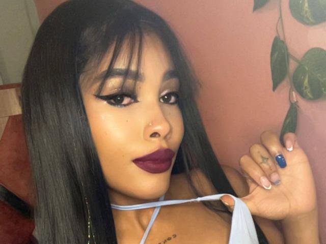 LexieBloom, con capelli neri e lisci, rossetto viola e eyeliner nero, tiene una tracolla blu, mostrando un piccolo tatuaggio sul petto, e ha un piercing al naso..