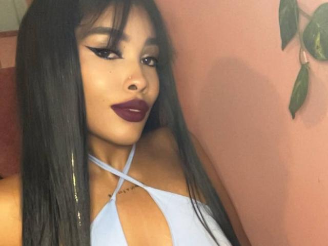 LexieBloom, con cabello negro largo, delineador de ojos oscuro y lápiz labial morado, vestida con un top de tirantes azul claro, que muestra su pecho y un pequeño tatuaje cerca de la clavícula..
