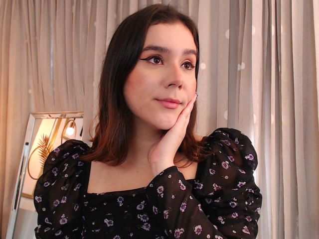 Deusa com cabelo castanho, lábios rosados e delineador alado, tocando sua bochecha com um leve sorriso. Ela está vestindo uma blusa preta com estampa floral e mangas bufantes..