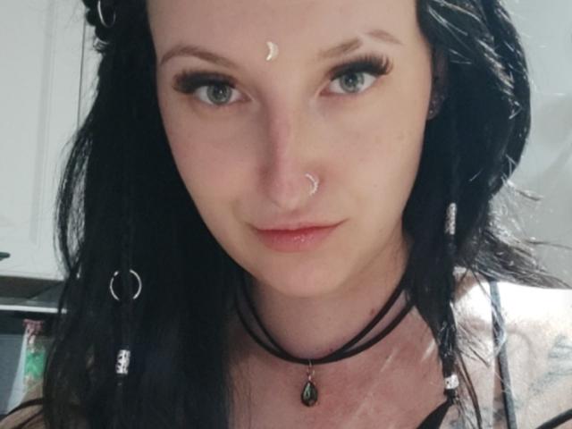 GoddessMoonx has pale skin, black hair, green eyes, and a crescent moon forehead tattoo. She's wearing a black choker with a teardrop pendant and a nose ring.