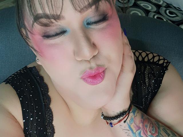 GiiannaMorrys with pink blush, blue eyeshadow, pouty pink lips, black lace top, tattooed arm, and nose ring, resting hand on cheek, short brown hair, gray couch background.