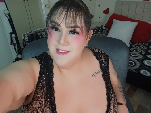 GiiannaMorrys with pink eyeshadow, black lace top, showing large tits, tattoo on right shoulder, smiling, dark hair, bangs, black chair, pink lipstick, and cute earrings.