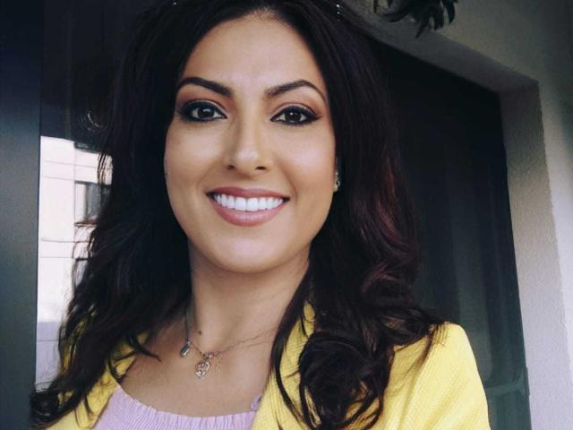 LisaCroftt has dark wavy hair, tan skin, and a big smile with white teeth. She's wearing a yellow cardigan over a pink top, and has subtle makeup.