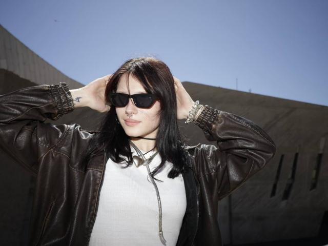 Bridget Norbury, con cabello castaño oscuro, gafas de sol negras, camisa blanca y chaqueta de cuero marrón, con las manos en el pelo, cielo azul claro, luz solar, informal, segura de sí misma, atractiva..