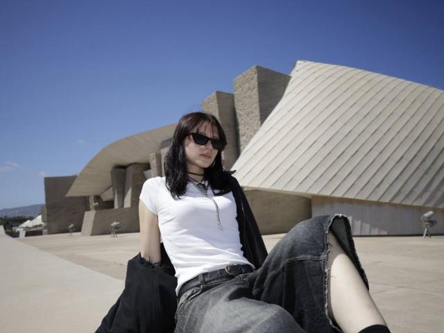 Bridget Norbury, con i capelli scuri, gli occhiali da sole, una maglietta bianca, una giacca nera e jeans strappati, seduta con una gamba sollevata, che mostra il sedere, di fronte a un edificio moderno..