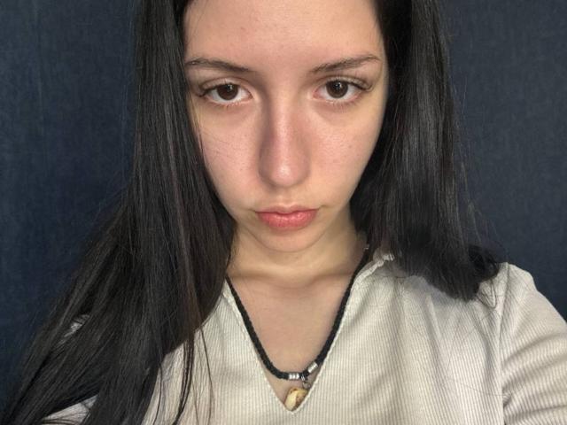 BethelVanhamme tiene el pelo largo y negro, ojos marrones y una expresión neutra. Lleva una camiseta blanca con textura y un collar negro con un pequeño colgante de madera..