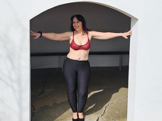 Dirty-Diva-hot with red bra, black textured pants, and black flats, standing with arms out, smiling, wearing a black wristwatch, under an archway, sunlight casting shadows.