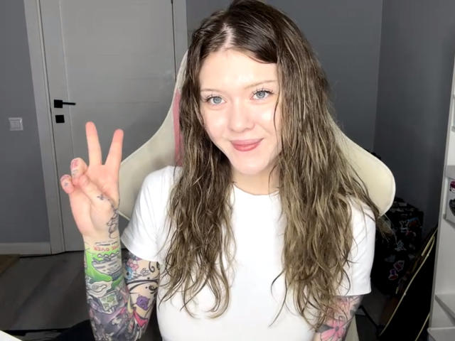 MeliaRinaldo with wet brown hair, white tee, tattooed arm, pinkie and middle finger up, smiling, blue eyes, fair skin, sitting in white chair, showing off her ass.