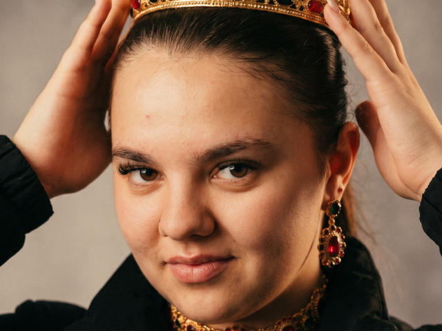 DouceLouise with her hands on a gold crown, wearing red and green earrings, black jacket, and gold necklace, smiling with brown eyes and subtle makeup.