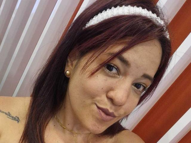LiaRamonaV has red hair, a white headband, small earrings, a gold necklace, and a tattoo on her left shoulder. She's smiling with her pouty lips.
