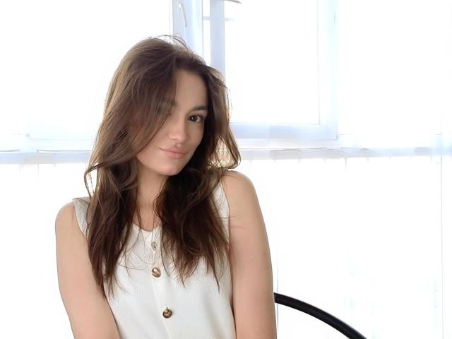 ShayneIwaoka with long brown hair, small tits, wearing a sleeveless white button-up, smiling softly, standing near a bright window, looking cute and relaxed.
