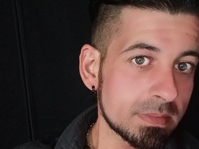 RiccoJohnson has a short, dark beard, a black ear gauge, and gray eyes, with his hair styled up and a slight smile. He's wearing a dark shirt.