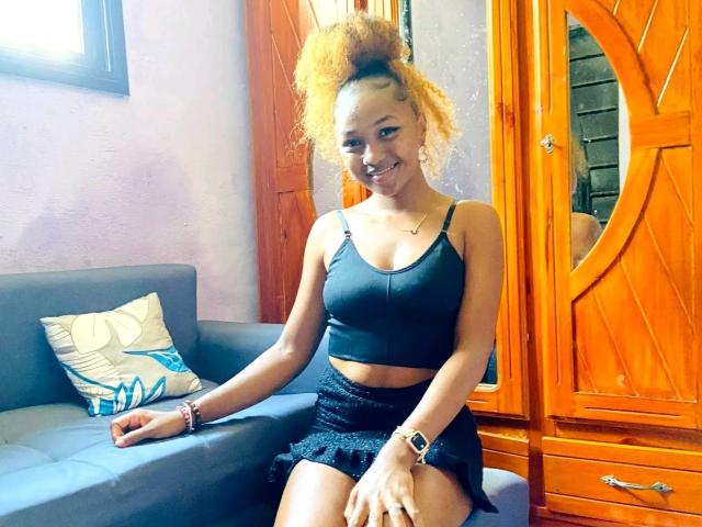 Jennitah, com cabelo loiro cacheado, top de ginástica preto, mostrando seus seios, e saia preta, sentada em um sofá cinza, sorrindo, armário de madeira ao fundo, relógio no pulso direito..