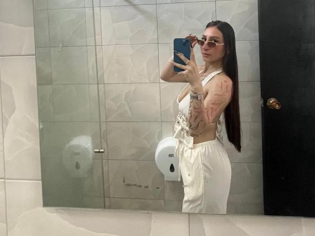 MaddyCarina with long brown hair, wearing white pants and a sleeveless top, showing tattoos on her arm and side, taking a mirror selfie with a blue phone.