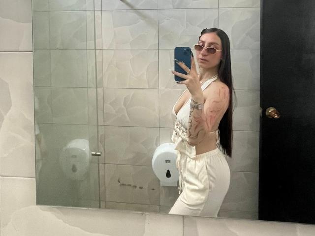 MaddyCarina with long dark hair, wearing sunglasses, a white side-tie top, and white pants, showing tattoos on her arm, taking a mirror selfie in a bathroom.