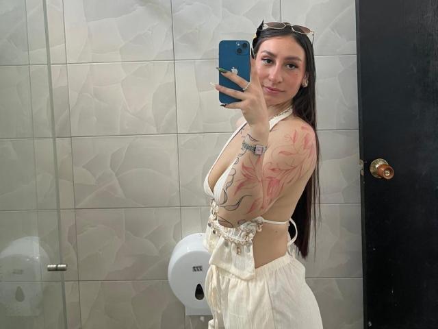 MaddyCarina with tattoos on her arm and side, wearing a white halter top and white skirt, taking a mirror selfie in a bathroom, showing her ass and small tits.