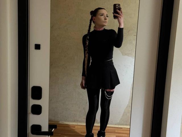AngiePlassmeyer with braided hair, wearing a black long-sleeve top, black skirt, and black leggings, taking a mirror selfie, showing off her slim, cute ass.