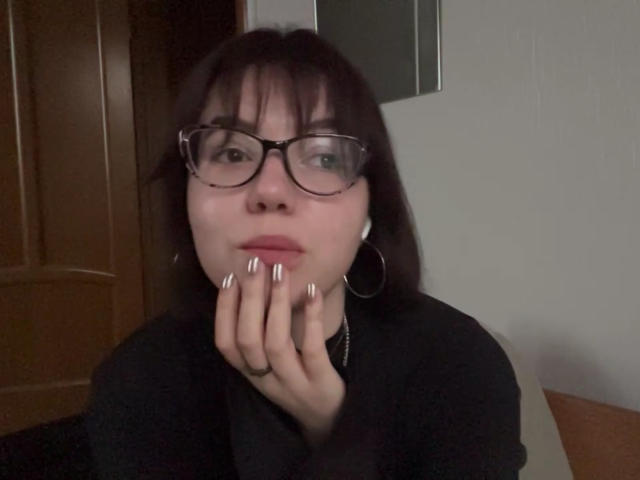 Carmon Yorton, com cabelo castanho escuro, óculos e unhas prateadas, apoiando o queixo na mão, vestindo uma camisa preta e brincos de argola, olhando para a câmera..