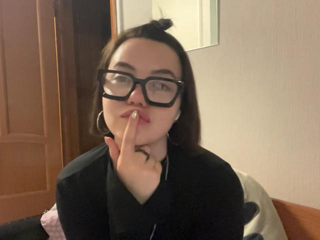 CarmonYorton con gafas negras, cabello castaño recogido en un moño, camisa negra, dedo tocando los labios, lápiz labial rosa, aros grandes, sentada en una cama, mirando a la cámara..