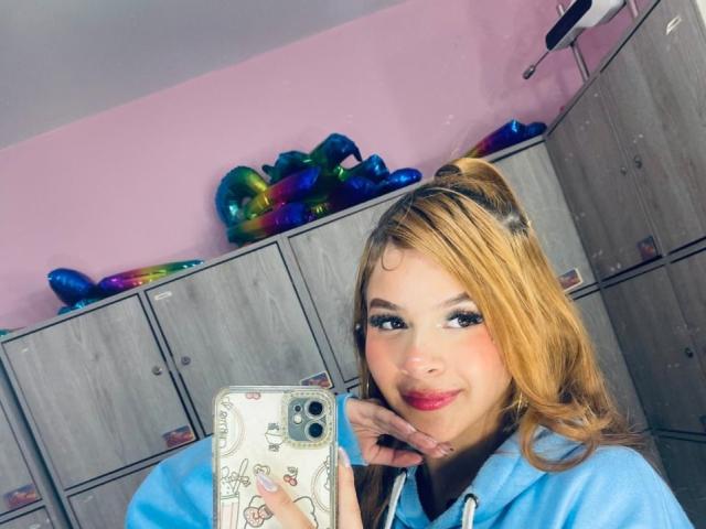 MadisonNoir con i capelli biondi raccolti in una coda di cavallo, indossa una felpa blu, tiene in mano un telefono, sorride, ha il rossetto rosa, si trova in una sala spogliatoio con palloncini colorati e armadietti di legno, ha la mano destra sul viso, scatta un selfie sexy..