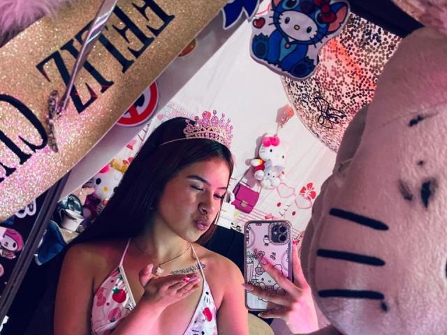 MadisonNoir, con un seno di media grandezza, indossa un top da bikini rosa a forma di cuore, una corona e ha un'espressione seria, mentre si scatta un selfie davanti a uno specchio decorato con immagini di Hello Kitty e decorazioni scintillanti..