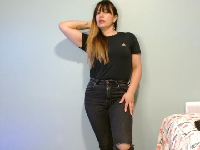 Roxanne Blair, con cabello largo y castaño, camiseta negra y jeans oscuros rotos, de pie frente a una pared azul claro, con la mano en el cabello, mirando a la cámara, tiene un tatuaje en el antebrazo izquierdo..