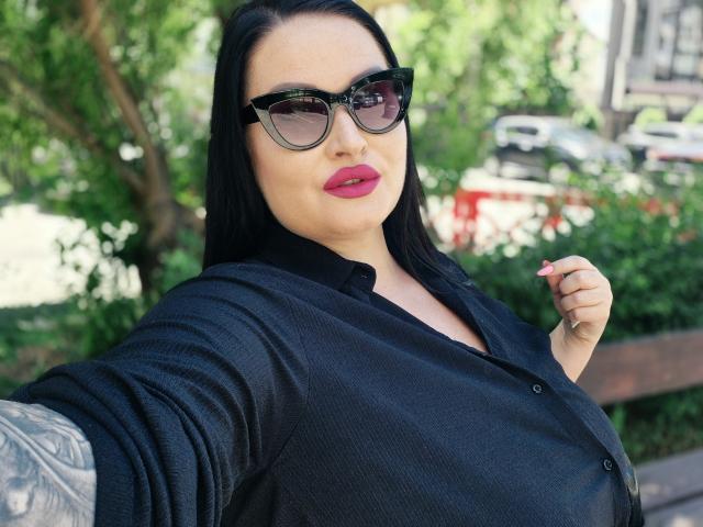 GeorgiaLerox with black hair, wearing black shirt, black sunglasses, and pink lipstick, showing tattooed arm, holding shirt, outside, sunny. Boobs visible under shirt.