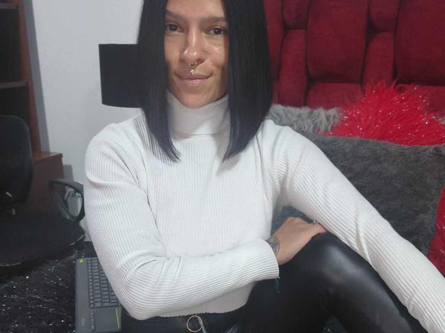 Amelie Blake, con capelli neri e lisci, maglia a coste bianca a collo alto, pantaloni in pelle nera, piercing al naso, seduta, sorridente, con un portatile su una scrivania nera, una sedia rossa e grigia sullo sfondo..