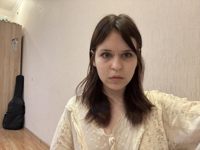LiliaDrube with a serious face, wearing a cream lace blouse, brown shoulder-length hair, and a black guitar against a beige wall in a wooden-floored room.