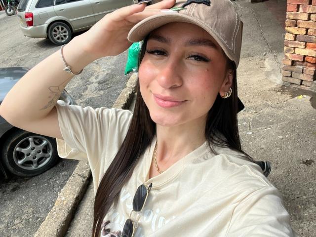 MaddyCarina with long dark hair, wearing a beige cap, beige tee, and gold earrings, holding sunglasses, smiling, hand up, tattoo on left arm, pinkish skin, street background.