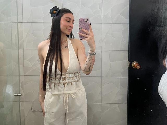 MaddyCarina with long black hair, white halter top showing big tits, white pants, black flower hair accessory, tattoos, holding phone, mirror selfie, smiling.