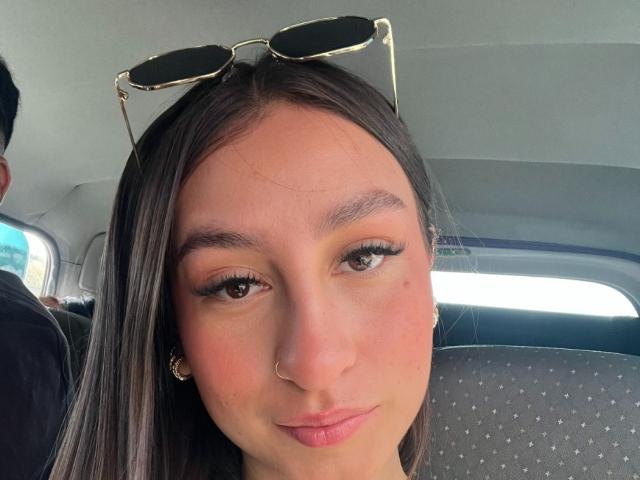 MaddyCarina's face in a selfie, showing brown eyes, full lips, and a small nose piercing. She has long brown hair, wears sunglasses on her head, and has a light skin tone.