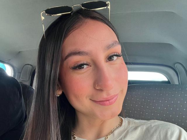 MaddyCarina with long black hair, green eyes, and a small nose ring, wearing sunglasses on her head, smiling in a car, white shirt, subtle makeup, and a gold necklace.