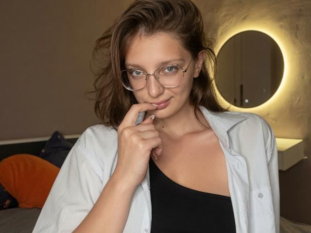 Stacey Averbach met een bril, bruin golvend haar, een wit shirt, een zwart tanktopje, die een vinger bijt, een schattende glimlach, zachte belichting, een ronde spiegel op de achtergrond, subtiele make-up, middelgrote borsten..