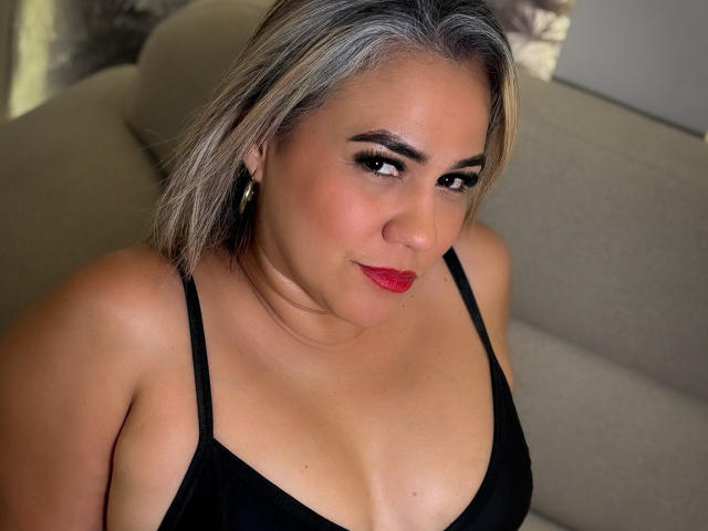 CamilaNoer with blonde hair, red lipstick, and black top, showing her tits, sitting on a beige couch, looking at the camera with a slight smile.