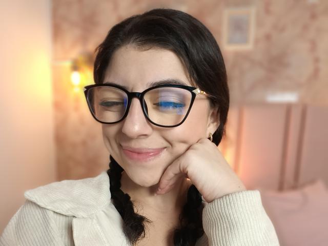 Anntonella Watson mit schwarzen Brillen, blauem Augen-Make-up, schwarzen Zöpfen, einem weißen Pullover, lächelnd, mit der Hand unter dem Kinn, sanfter rosa Beleuchtung, leicht zwinkernd, süß, entspannt, Schlafzimmerszenario..