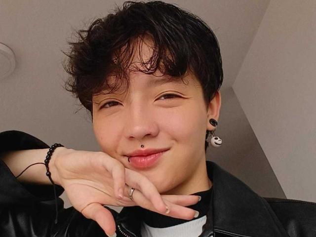 A LissyLuxe tem cabelo preto curto e bagunçado, um piercing no nariz e brincos de argola pretos. Ela está vestindo uma jaqueta de couro preta, sorrindo com o dedo perto da boca..