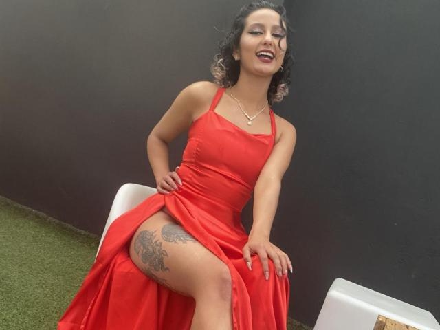 Flora Saenz, com cabelo preto encaracolado, vestindo um vestido vermelho que expõe a coxa tatuada, sentada em uma cadeira branca, sorrindo, com as mãos no vestido, mostrando a vulva através da fenda alta do vestido..