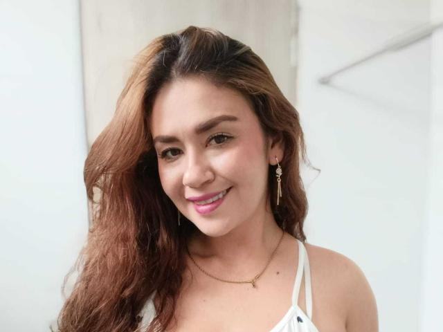 TheresaLarson has wavy brown hair, pink lipstick, and a slight smile. She's wearing a white halter top, gold necklace, and dangling earrings, looking cute and sexy.