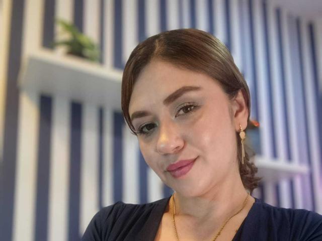 TheresaLarson has fair skin, brown hair in a ponytail, green eye makeup, pink lipstick, and gold earrings. She's wearing a black top with a gold necklace.