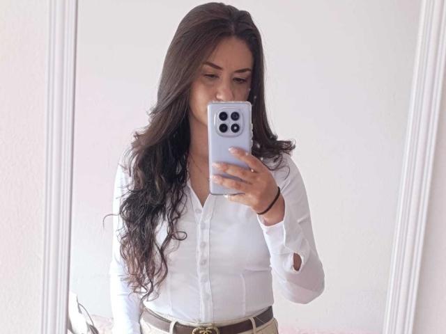 TheresaLarson with long wavy brown hair, white button-up shirt, and brown belt, taking a mirror selfie with her iPhone, showing her tits and a cute smile.