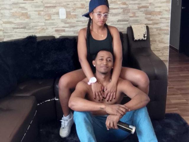 LilithAndAsmodeo with small tits and dark ass sits on a shirtless guy's lap, wearing a black tank top and blue cap, holding a remote in a living room.