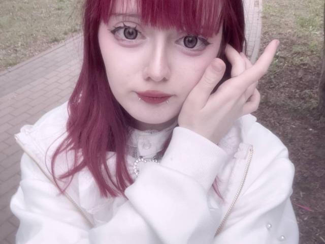 Robin Krapp mit rosa-lila Haaren, heller Haut und Eyeliner, die sich eine Hand vor das Gesicht hält, trägt eine weiße Jacke mit Knöpfen und eine silberne Halskette, auf einem Selfie..
