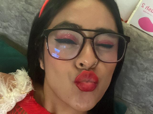 StellaBeckker, con unos labios rojos prominentes, párpados hinchados y gafas de montura negra gruesa, dando un beso con puchero. Tiene el pelo oscuro, un piercing en la nariz y una diadema roja..