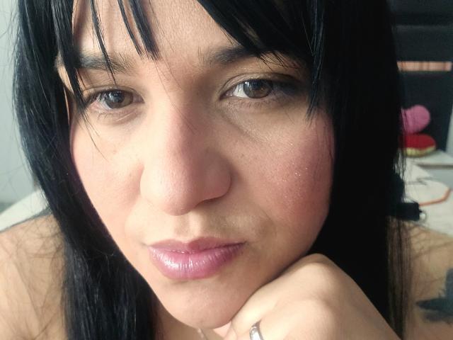 Il viso di Viviana Thompson, capelli neri, occhi marroni, labbra rosa, leggera abbronzatura, lentiggini visibili, una mano appoggiata sul mento con un anello, luce soffusa, espressione casual, naso leggermente lucido..
