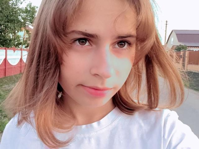 MireilleSpotwood has brown hair, brown eyes, and a light skin tone with a slight blueish shadow on her right cheek. She's wearing a white shirt, looking at the camera.