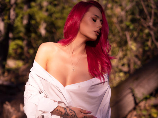 KaylieRosse with bright pink hair, white shirt off shoulders, exposing her tits, tattoo on right hand, necklace, eyes closed, sunlight, forest background.